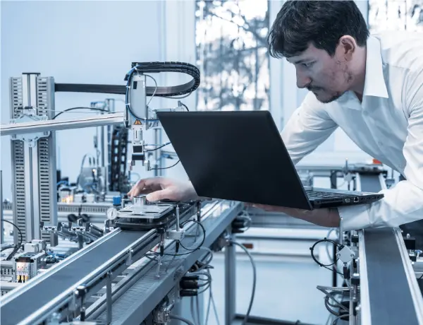 Ingeniero con laptop supervisando línea de producción automatizada mediante programación en PLC.
